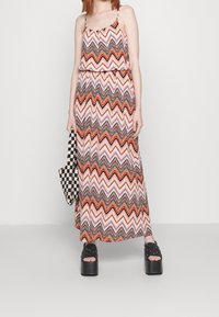 Vrouw draagt een mouwloze maxi-jurk met zigzagpatroon in oranje, zwart en wit, houdt een ruitvormige tas vast en draagt zwarte platformsandalen.