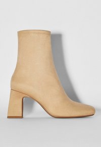 Stivaletto in suede beige con punta quadrata e tacchi grossi e rettangolari. Il collo presenta dettagli cuciti lungo il bordo superiore.