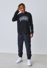 Sort langærmet skjorte med "BAKER" grafik; mørke cargobukser; hvide sneakers; fletninger i håret; neutral baggrund.