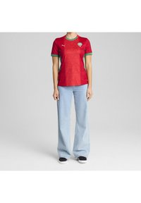 Osoba ubrana w czerwono-zielony krótki rękawowy top sportowy i jasnoniebieskie szerokie dżinsy stoi na jednolitym szarym tle.