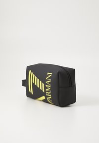 Bolsa de cosméticos preta com design gráfico amarelo e logótipo, feita de material texturizado, possui fecho de zip e uma alça lateral. Forma retangular.