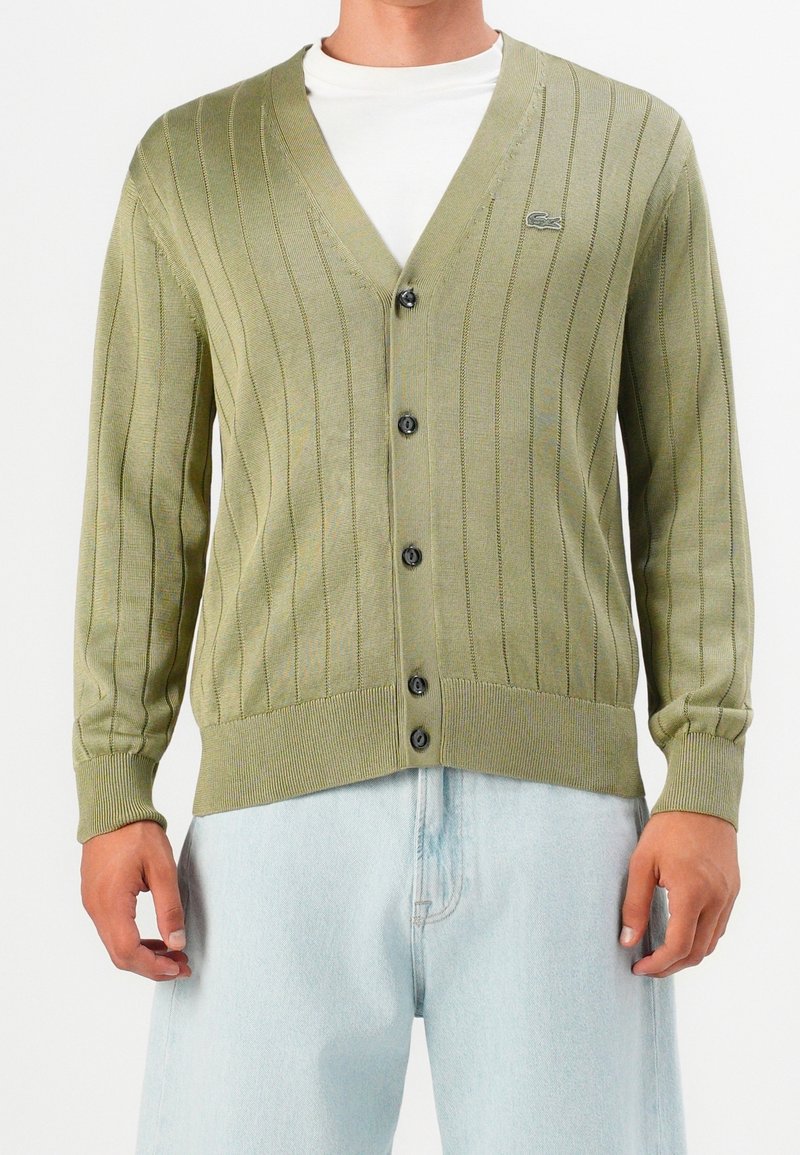 Homme portant un cardigan côtelé vert clair avec des boutons noirs par-dessus une chemise blanche et un jean bleu clair, visible du cou jusqu'aux hanches.