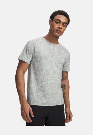 Mann trägt ein hellgraues, kurzärmeliges, gemustertes T-Shirt und schwarze Hose, steht mit neutralem Gesichtsausdruck und hat ein Tattoo am linken Unterarm.