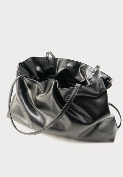 Grand sac fourre-tout en cuir noir avec deux fines bretelles et une forme décontractée et souple sur un fond uni.