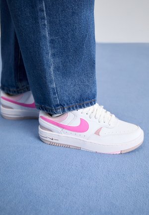 Białe trampki z różowym logo Swoosh noszone z niebieskimi dżinsami, stojące na jasnoniebieskim wykładzinowym podłodze.