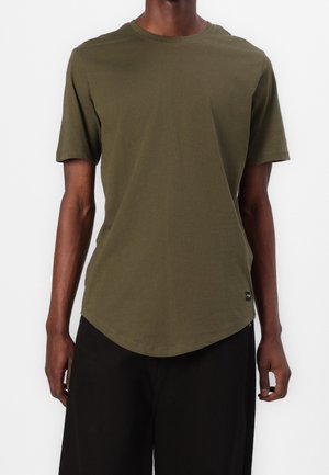 Mann trägt ein schlichtes olivgrünes kurzärmliges T-Shirt und schwarze Hose, steht vor einem weißen Hintergrund.