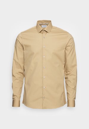 Beige langærmet skjorte med knapper, klassisk krave og hvide knapper, vist på en ensfarvet lys baggrund.