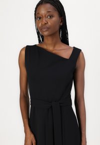 Robe noire sans manches avec un décolleté asymétrique et une taille nouée. Fabriquée dans un tissu lisse et léger, avec un design simple.