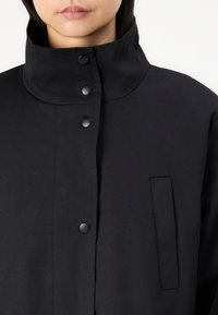 Veste noire avec un col montant, des boutons-pression et une poche latérale. Fabriquée dans un tissu texturé avec une finition lisse.