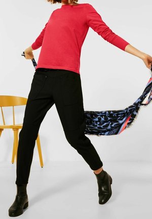 Červené tričko s dlouhým rukávem, černé kalhoty na míru s kapsami a černé kotníkové boty. V jedné ruce drží barevnou šálu na jednoduchém pozadí.