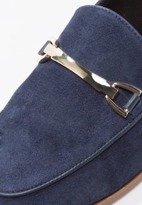 Mocassins en daim bleu marine avec un accent métallique doré et des coutures le long du bord. Semelle plate avec une texture lisse.