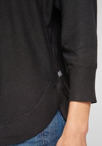 Czarny sweter z dzianiny z okrągłym dołem, krótkimi rękawami z mankietami oraz małą naszywką przy szwie, zaprezentowany w zestawieniu z dżinsami.