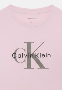 Svetloružové bavlnené tričko s veľkým sivým logom "CK" a čiernym textom "Calvin Klein". Okrúhly výstrih so šitými hranami.