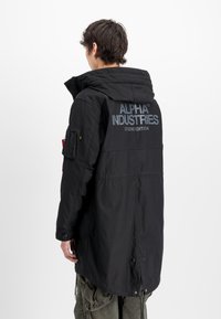 Parka negra con capucha, que presenta un gran logo de "ALPHA INDUSTRIES" en la parte posterior, múltiples bolsillos y un dobladillo con cordón.