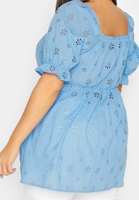 Niebieska bawełniana bluzka z kwadratowym dekoltem, bufkowatymi rękawami i wzorem kwiatowych ażurowych zdobień. Marszczona talia dodaje kształtu.