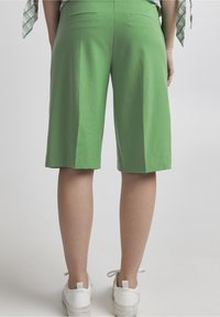 Osoba stojící v zelených šortkách po kolena, bílých teniskách a zkráceném kostkovaném topu, zobrazená zezadu na jednobarevném pozadí.