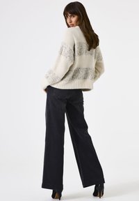 Eine Frau trägt einen cremefarbenen Strickpullover mit silbernen Pailletten und schwarze weit geschnittene Hosen. Sie steht mit den Händen in den Taschen vor einem weißen Hintergrund.