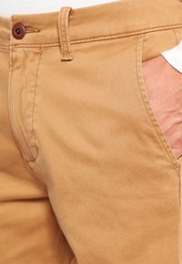 Béžové bavlněné chinos s plochým předním dílem, bočními kapsami a hnědým knoflíkem na zavírání v pase. Hladká textura, střih s rovnou nohavicí.