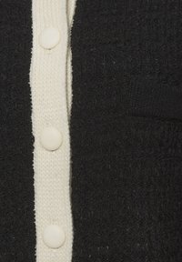 Juodos ir baltos spalvų megztinis su tekstūruotu juodu kūnu, lygiu baltu apvadu, paprastais apvaliais mygtukais ir maža kišene.