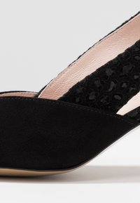Černé semišové wedge boty s texturovanými černými látkovými akcenty, otevřenými stranami a béžovou podšívkou. Zakřivený tvar a špičatý design špičky.