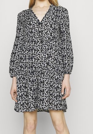 Nainen, joka pukeutunut mustavalkoiseen kuvioituun pitkähihainen mekkoon, jossa on V-pääntie ja hieman liehukehelmainen hame.