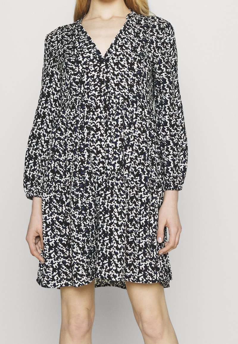 Femme portant une robe à manches longues à motifs noir et blanc, avec un col en V et une jupe légèrement évasée.