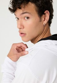 Sudadera blanca con interior negro, que cuenta con un bolsillo frontal, un ligero brillo y cordones. Se pueden ver rizos en el cabello y un pendiente de aro plateado.