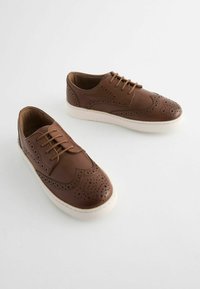 Hnědé kožené boty s brogue detaily, šněrovacím designem a bílými gumovými podešvemi. Textura je hladká s perforovanými akcenty.