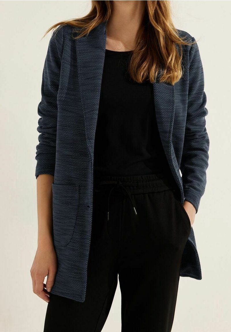 Femme portant un blazer bleu foncé texturé ouvert sur un haut noir et un pantalon noir à cordon, debout avec une main dans la poche.