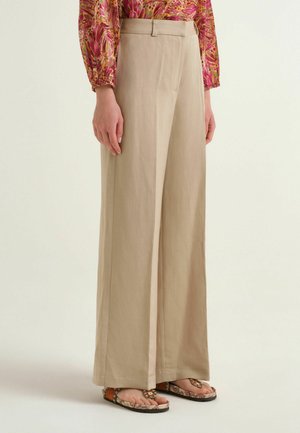 Pantaloni beige a gamba larga abbinati a sandali con motivo e ad una camicetta floreale colorata a maniche lunghe su una persona in piedi.