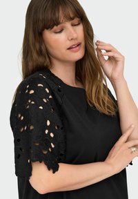 Woman with long brown hair wearing a black top with floral lace sleeves, gently touching her hair and showing a ring on her finger.