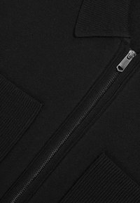 Juodas prakaitavimas su užtrauktuku, pasižymintis lygiu megztu tekstūru, raukšlėtomis detalėmis ir sidabriniu užtrauktuku. Dizainas apima dvi priekinės kišenes.