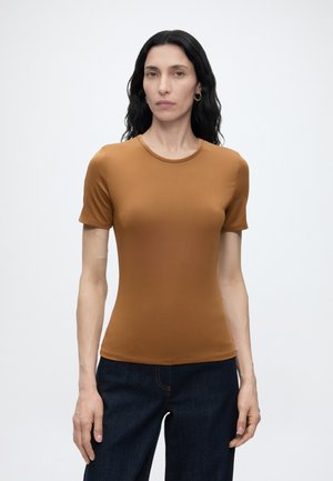 Mujer con cabello largo y negro que lleva una camiseta marrón ajustada de manga corta y vaqueros azul oscuro sobre un fondo blanco liso.