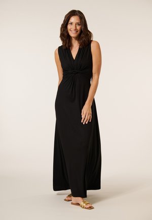 Glunderende vrouw in een mouwloze zwarte maxi-jurk met gedraaide voorkant, met gouden sandalen, tegen een lichte beige achtergrond.