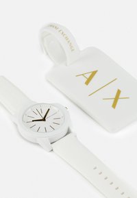 Orologio analogico bianco con cassa rotonda, marcatori delle ore dorati e cinturino in silicone. Include un'etichetta bianca con "A/X" in lettering dorato.