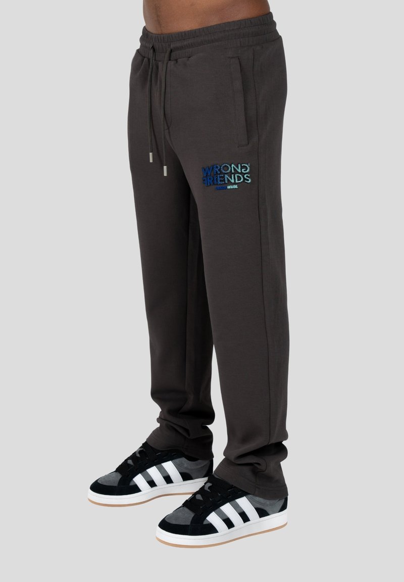 Graue Sweatpants mit elastischem Bund und Kordelzug, mit dem gestickten Text "WRONG FRIENDS" in buntem Garn. Schwarz-weiße Sneakers.