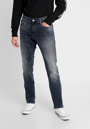 Hombre con vaqueros azul oscuro desteñidos, camiseta negra de manga larga y zapatillas blancas, de pie con una mano en el bolsillo.