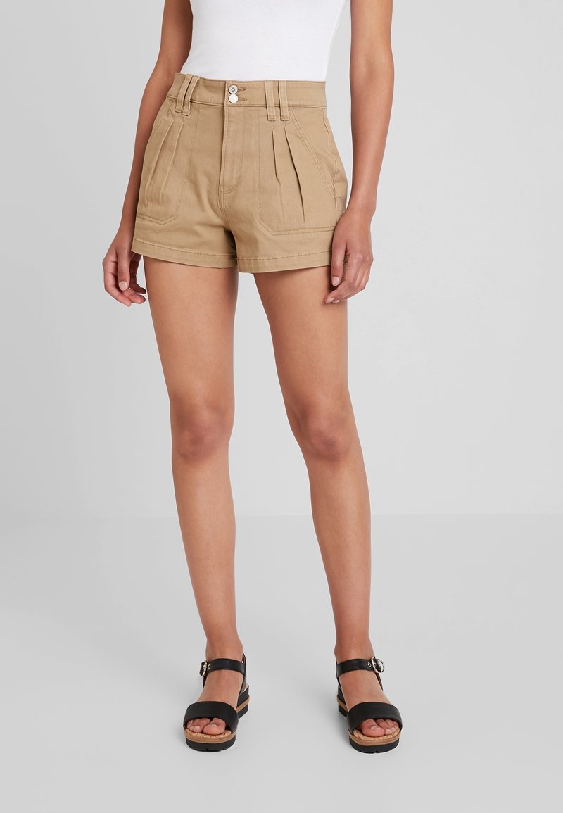 Tan high-waisted shorts with pleats, two front pockets, and a zipper fly. Paired with black platform sandals. Smooth cotton material.