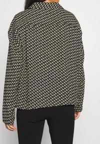 Schwarz-cremefarbenes Shirt mit Zickzackmuster, Kragen, langen Ärmeln und lockerem Sitz, das eine glatte Textur zeigt.