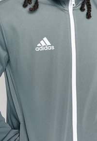 Grå atletjakke i struktureret stof. Har en hvid lynlås og et broderet hvidt Adidas-logo på venstre bryst.