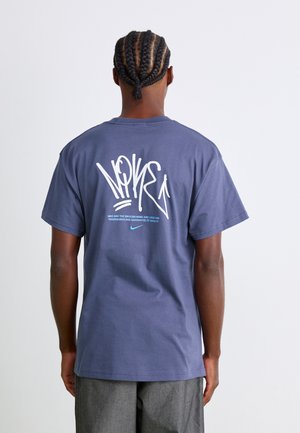 Person med fletninger iført en blå Nike t-shirt med et stort hvidt grafisk motiv og tekst på ryggen, parret med grå bukser.