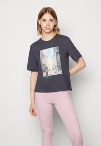 T-shirt de algodão azul marinho com gráfico de paisagem urbana. Modelo usa com leggings de riscas rosa claro. Mangas curtas, corte descontraído.
