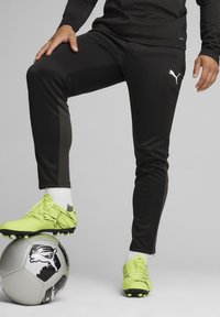 Schwarze Puma-Sporthosen mit dunkelgrauen Einsätzen, kombiniert mit leuchtend gelben Fußballschuhen, die auf einem silbernen Fußball mit schwarzen Akzenten ruhen.