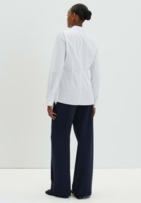 Weißes Langarmhemd mit Button-down-Kragen, kombiniert mit navyblauen weiten Hosen. Glatte Stoffoberfläche mit sichtbaren Nähten und Rückendetails.