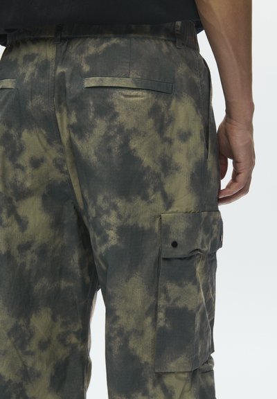 Πίσω όψη ατόμου που φοράει cargo παντελόνια με μοτίβο tie-dye σε πράσινο και μαύρο, με τσέπες με καπάκι, στέκεται μπροστά σε λευκό φόντο.
