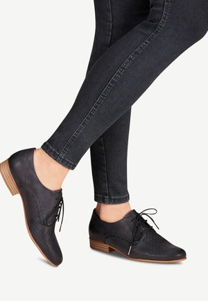 Voeten met zwarte, getextureerde leren veterschoenen in oxfordstijl met houten hakken, gecombineerd met donkergrijze skinny jeans tegen een witte achtergrond.