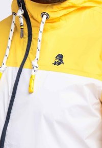 Chaqueta cortavientos amarilla y blanca con un logo negro, que cuenta con una capucha ajustable y detalles en el cierre de contraste. Tejido texturizado.
