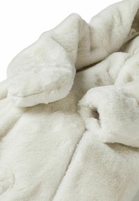 Chaqueta de piel sintética blanca con textura suave, mangas de gran tamaño y bolsillos laterales, que cuenta con un cierre de cremallera y un forro suave.
