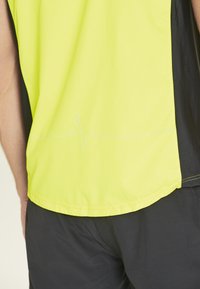 Leuchtend gelbes Sportshirt mit schwarzen Seitenpaneelen, das am Saum ein Herzschlaglinien-Grafik in Hellblau aufweist.