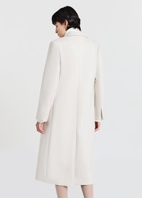 Cappotto lungo color crema con collo a revers, texture liscia e spacchi laterali. Presenta una silhouette aderente e maniche lunghe.